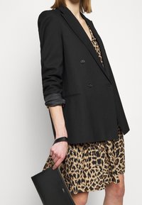 Personne portant un blazer noir croisé sur une robe à imprimé léopard, tenant une pochette noire avec une lanière pour poignet.