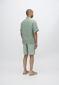 Light green linen shirt with short sleeves, paired with matching textured shorts. Model wearing tan slide sandals.