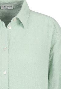 Hellgrünes, strukturiertes Hemd mit einem Button-Down-Kragen, das kleine erhabene Punkte aufweist und perlenartige Knöpfe entlang der Vorderleiste hat.