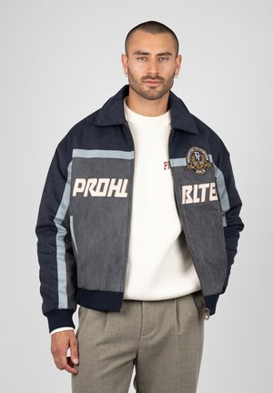 Man with short hair wearing navy and gray zip-up jacket with "PROHL BLTE" text and a patch, white sweater, and beige pants, standing against plain background.