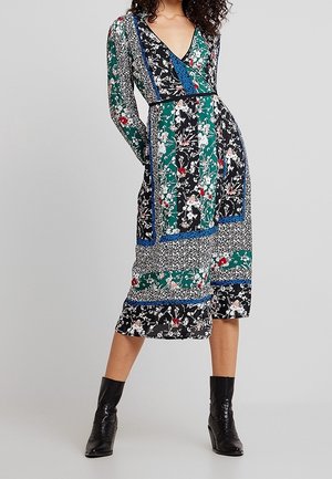 Vrouw draagt een knielange jurk met bloemenpatchwork, lange mouwen en zwarte enkellaarsjes met hak, staand tegen een effen achtergrond.