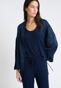 Granatowa kurtka w stylu kimono z zielonym abstrakcyjnym wzorem, z przezroczystego materiału, luźny fason, dobrana z granatową bluzką na ramiączkach i pasującymi spodniami, casualowy design.