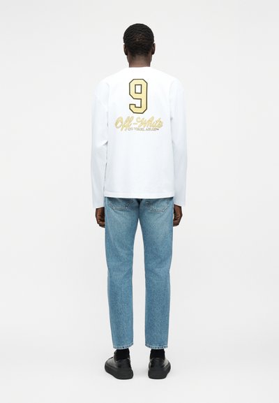 Άνδρας που φοράει λευκό πουκάμισο με μακριά μανίκια, με μεγάλο αριθμό 9 και το κείμενο "Off-White c/o Virgil Abloh" στην πλάτη, μπλε τζιν και μαύρα παπούτσια.