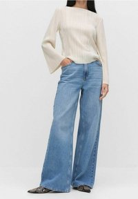 Světle béžové tričko s dlouhým rukávem a svislými záhyby, spojené s modrými džínami s širokými nohavicemi. Oblečení má volný střih a rozšířené rukávy.