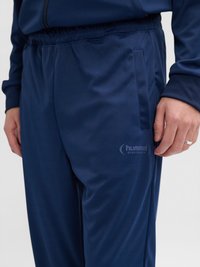 Nærbillede af en person iført marineblå sportbukser med sidelommer og et "hummel sportswear" logo på låret.