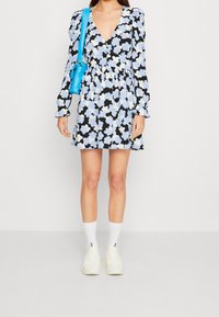 Blommig klänning med djup V-ringning, svart bakgrund, vita och blå blommor, långa ärmar med volanger. Matchad med en blå axelväska och vita sneakers.