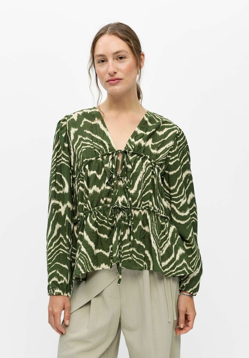 Femme portant un chemisier à manches longues vert et blanc à motifs avec des détails noués et un pantalon ample beige clair sur un fond uni.