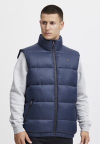 Laikku sininen pufferi liivi korkealla kauluksella, edessä vetoketju ja sivutaskut; siinä on tikattu kuvio ja kiiltävä kangas; päällä harmaa pitkähihainen paita.