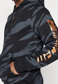 Zwarte hoodie met grijze abstracte patronen. Heeft een voorvak, trekkoorden en bronzen logo-accenten op de mouw.