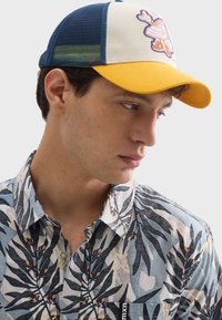 Modrá a žltá baseballová čiapka s mriežkovanými stranami, zdobená karikatúrou. Nositeľ má na sebe košeľu s kvetinovým vzorom, tmavými listami a svetlými akcentmi.