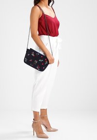 Tmavě modrá taška přes rameno s vyšívaným motivem třešní, obdélníkového tvaru, s řetízkovým popruhem a kovovým zapínáním. Nošena s bordó topem a bílými kalhotami.