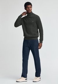 Dunkelgrüner Strickpullover mit Rollkragen, gerippten Bündchen und Saum, kombiniert mit dunklen Jeans und hellen Sneakern.
