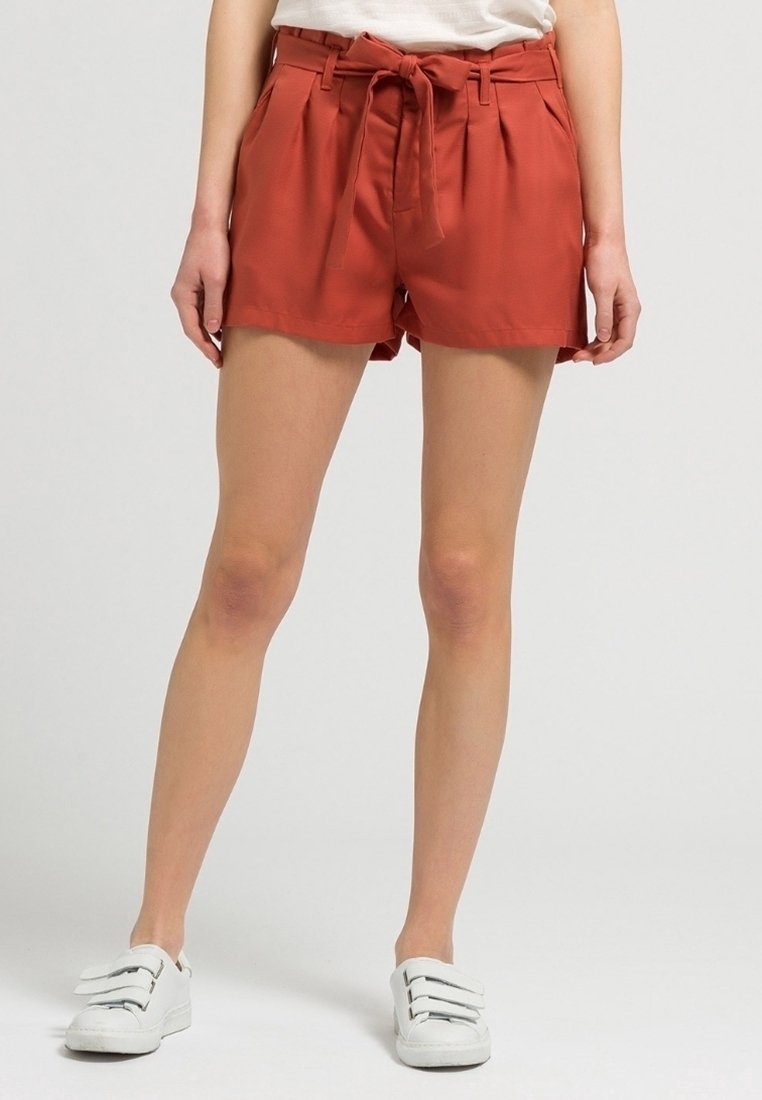 Femme portant un short taille haute couleur rouille avec lien à la taille, un haut blanc et des baskets blanches à scratch, debout devant un fond gris uni.