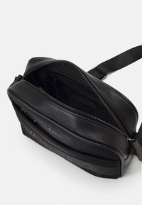 Černá kožená kabelka přes rameno se zipovým uzávěrem, s hladkou texturou, nastavitelným popruhem a decentním logem na přední straně.