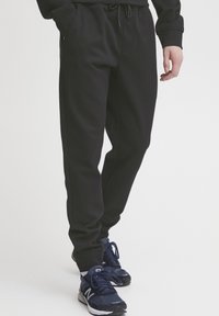 Pantalon de survêtement noir avec taille à cordon, poches latérales et poignets côtelés, associé à des chaussures de sport bleu foncé. Tissu lisse et léger.