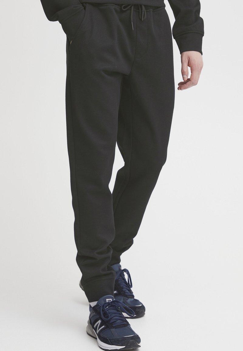 Pantalon de survêtement noir avec taille à cordon, poches latérales et poignets côtelés, associé à des chaussures de sport bleu foncé. Tissu lisse et léger.