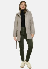 Cappotto grigio in misto lana con cappuccio, tasche frontali e vita con nastro, abbinato a un dolcevita nero, pantaloni verde oliva e sneaker beige.