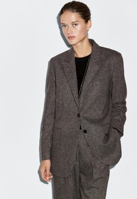 Donna che indossa un completo in lana grigia scura su misura con una maglia nera, in piedi con una mano in tasca contro uno sfondo chiaro e semplice.