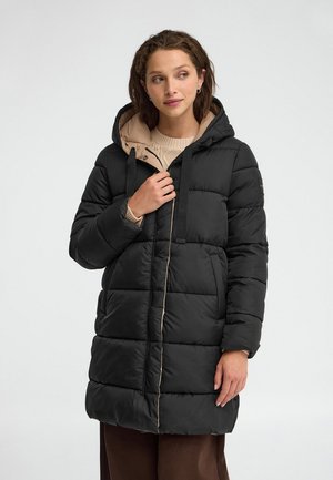 Donna che indossa un lungo piumino nero con cappuccio e fodera beige, in piedi davanti a uno sfondo chiaro e uniforme, che guarda di lato.