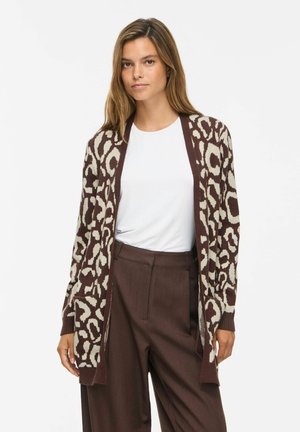 Kvinde iført en hvid t-shirt, brun mønstret åben cardigan og brune højtaljede bukser står mod en hvid baggrund.