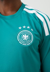 Türkisfarbenes Sporttrikot mit weißen Streifen auf den Schultern. Mit rundem, besticktem Abzeichen mit Adler und fünf Sternen darüber.