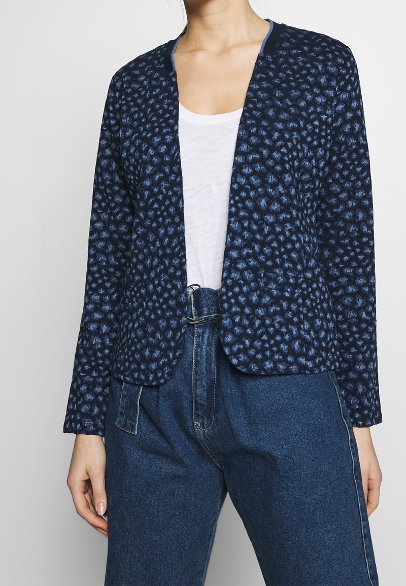 Cardigan color navy con un motivo testurizzato che presenta accenti di blu più chiaro, design aperto nella parte anteriore e maniche lunghe, abbinato a un top bianco e pantaloni in denim.