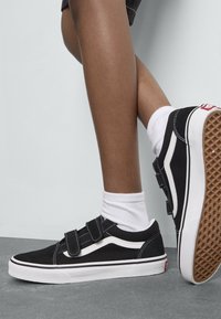 Black velcro sneakers with white accents, suede upper, rubber sole, and a textured gum outsole. Worn with white ankle socks.