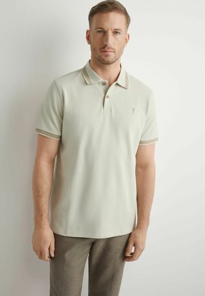 Mężczyzna w jasnobeżowej koszulce polo z paskowanym kołnierzykiem i brzegami rękawów, w brązowych spodniach, stojący na białym tle.