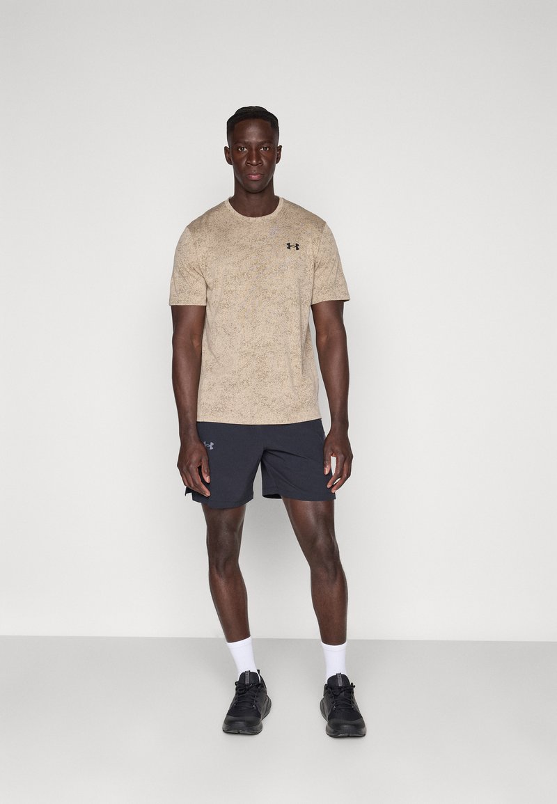 Hombre de pie vistiendo una camiseta beige de Under Armour, pantalones cortos negros, calcetines blancos y zapatillas negras contra un fondo blanco liso.