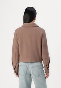 Bruine korte jas met een geknipte kraag en knoopmanchetten, gemaakt van een zachte stof. Gecombineerd met lichtblauwe denim jeans.
