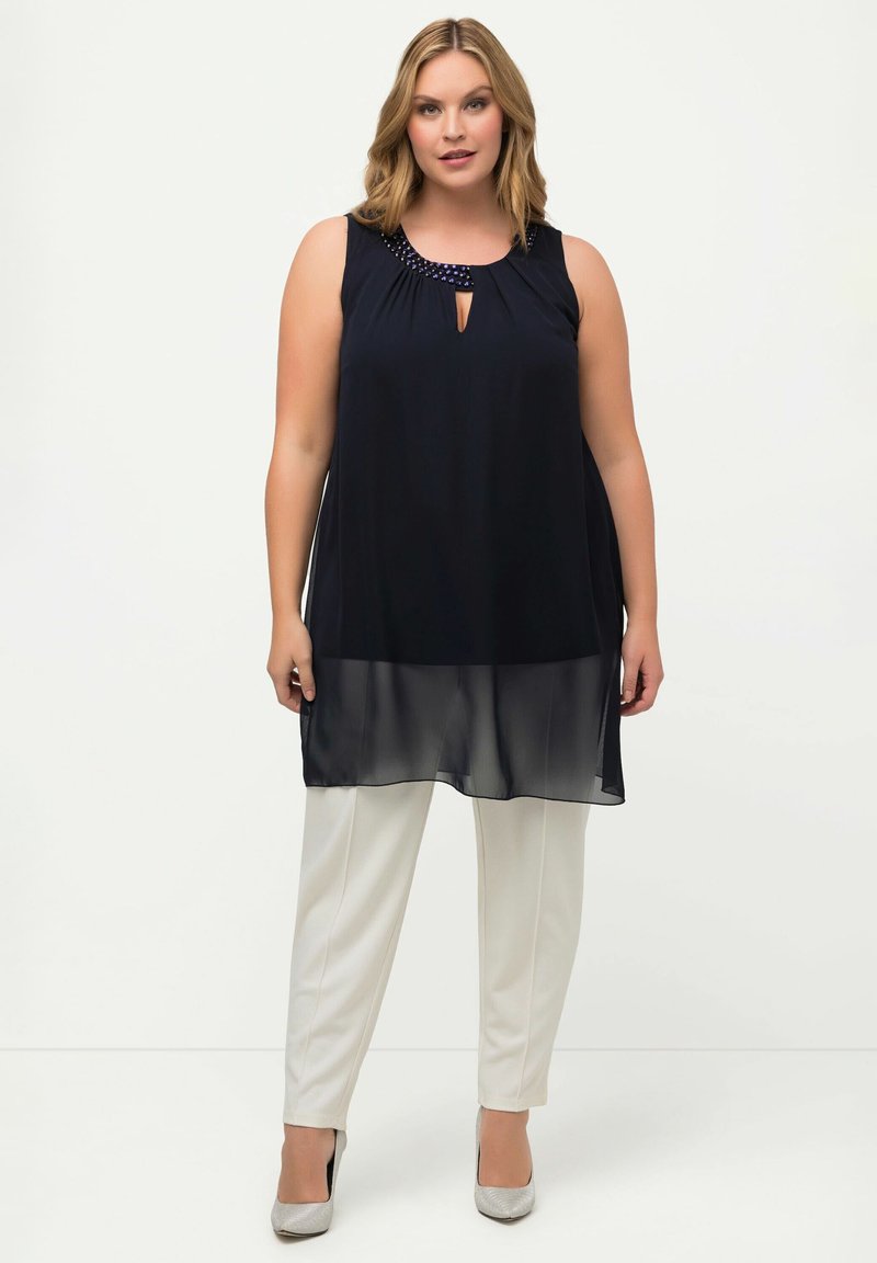 Top senza maniche blu navy con scollatura a chiave e dettagli di perline, abbinato a pantaloni bianchi su misura e tacchi argento, caratterizzato da una texture fluida.