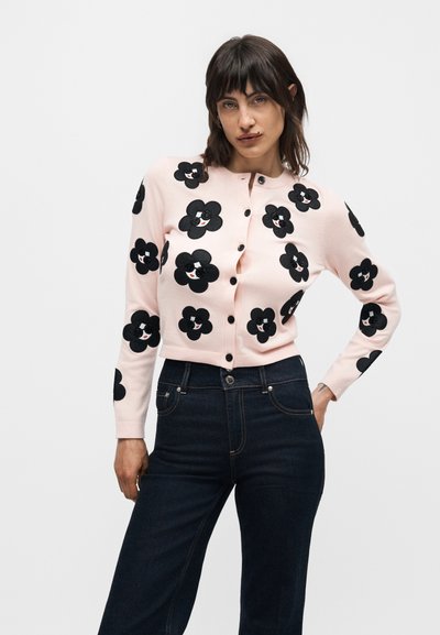 Γυναίκα που φοράει μια ανοιχτό ροζ ζακέτα με κουμπιά και μαύρα ανθισμένα μοτίβα, συνδυασμένη με σκουρόχρωμο ψηλόμεσο τζιν, ποζάροντας σε λευκό φόντο.
