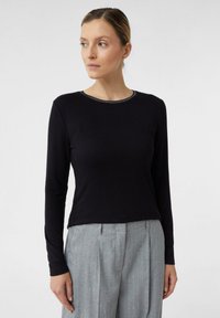 Schwarzes Langarmshirt aus weichem Material mit einem runden Ausschnitt und einer subtilen metallischen Einfassung. Kombiniert mit hellgrauen, gestreiften Hosen.