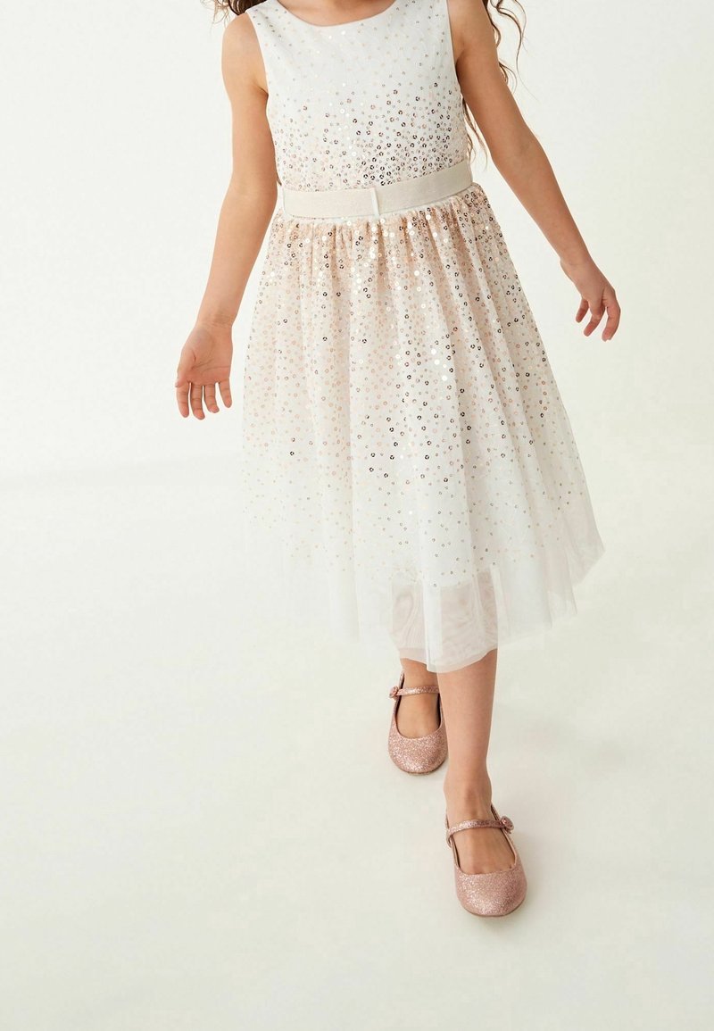 Fille portant une robe blanche sans manches ornée de sequins dorés et des chaussures Mary Jane roses pailletées assorties, marchant vers l'avant sur un fond uni.