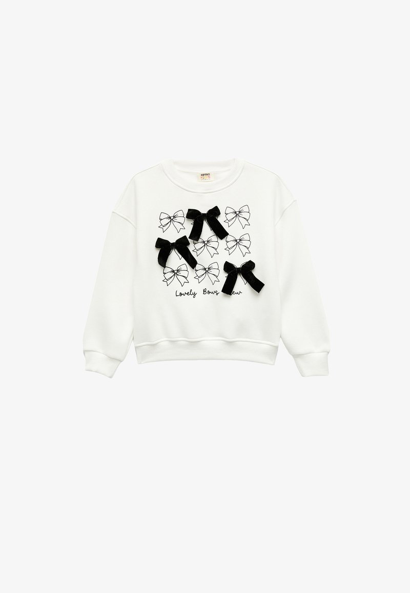 Sudadera blanca con un estampado de lazo en blanco y negro, acentuada con tres lazos de tela negra cosidos en el frente. Mangas largas, cuello redondo.