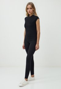 Una giovane donna sta in piedi con sicurezza indossando un top nero aderente e leggings, abbinati a sneakers bianche, su uno sfondo bianco semplice.
