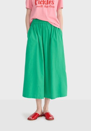 Žena v zelených voľných nohaviciach s širokými nohavicami, ružovom tričku s potlačou a červených sandáloch s otvorenou špičkou, stojaca s rukami v vreckách.