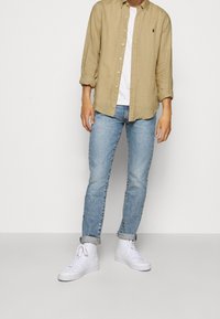 Khaki koszula z długim rękawem na białym t-shircie, w zestawieniu z jasnoniebieskimi dżinsami i białymi butami sportowymi za kostkę. Widoczne podwinięte rękawy i mankiety.