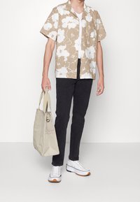 Camicia a maniche corte con motivo floreale beige e bianco, abbinata a jeans scuri e sneakers bianche. Una borsa a tracolla beige pende dal braccio.