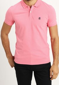 Růžová polo košile vyrobená z bavlny, s límečkem, krátkým rukávem a malým vyšitým logem na levé hrudi. Nohavice jsou tmavé.