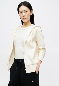 Šviesiai kreminės spalvos zip-up džemperis su reguliuojamu gobtuvu, minkšta tekstūra, derinamas su prigludusia balta ripsuota maike ir juodomis sportinėmis kelnėmis su auksiniu logotipu.