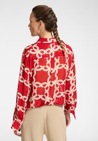 Blusa rossa con motivo a fantasia e disegni geometrici crema, maniche lunghe, colletto a bottoni e vestibilità morbida, mostrata di spalle.