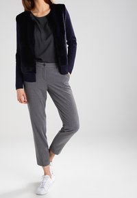 Veste en fausse fourrure bleu marine sur un chemisier gris, associée à un pantalon gris sur mesure et des baskets blanches. Tissu lisse, coupe moderne, style décontracté.