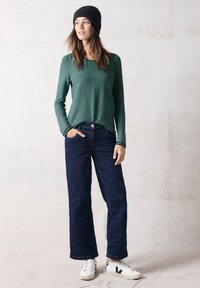 Grön räfflad långärmad topp, marinblå vida byxor och vita sneakers med svarta detaljer. Modellen poserar med en hand i fickan.
