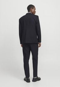 Dunkelblauer Anzug mit strukturierter Jacke und schmal zulaufenden Hosen, mit flacher Vorderseite, einem einzigen Rückenschlitz und glänzenden schwarzen Schuhen.