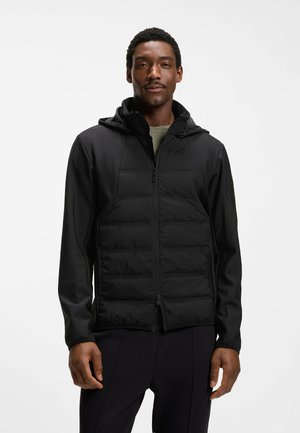 Hombre con chaqueta acolchada con capucha negra, con cremallera cerrada, de pie frente a un fondo blanco liso.