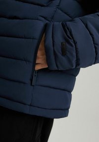 Granatowa kurtka puchowa z zamkiem błyskawicznym i pikowanym wzorem. Tkanina ma gładką teksturę o matowym wykończeniu i subtelnym detalu z logo.