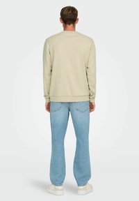 Helles beiges Sweatshirt mit Rundhalsausschnitt, gerippten Bündchen und Saum, kombiniert mit hellblauen Jeans in lockerer Passform und weißen Sneakers.