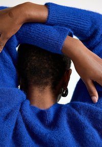 Královsky modrý pletený svetr se strukturovanou tkaninou, s žebrovanými manžetami a uvolněným střihem. Ruce spočívají na ramenou a odhalují jemný detail příze.