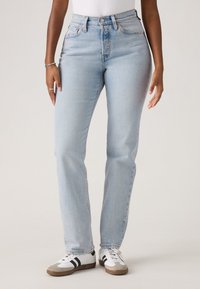 Persona che indossa jeans a gamba dritta azzurro chiaro e sneakers bianche con strisce nere, in piedi di fronte a uno sfondo chiaro e semplice.